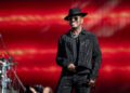 Ne-Yo in a black hat and sparkly outfit performs on stage with a red background.