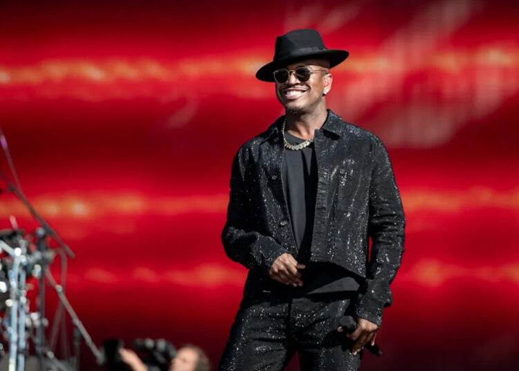 Ne-Yo in a black hat and sparkly outfit performs on stage with a red background.