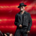 Ne-Yo in a black hat and sparkly outfit performs on stage with a red background.