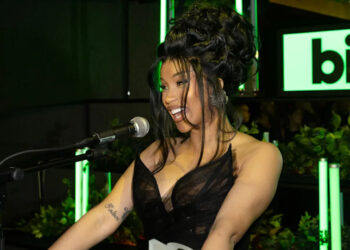 Cardi B, in a black dress, speaking into a microphone at a Billboard event, with green lights and plants in the background.