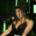 Cardi B, in a black dress, speaking into a microphone at a Billboard event, with green lights and plants in the background.