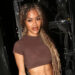 Teyana Taylor posing in a brown crop top, showcasing her toned abs, with long, wavy hair, standing in front of stage equipment.