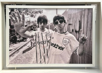 A black-and-white photo of Oasis band members Liam and Noel Gallagher wearing Manchester City football jerseys. Liam holds his hand up, palm facing out, while Noel holds his hand up with his index finger pointing to the sky.