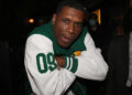 Jay Electronica in a green and white varsity jacket poses with arms crossed. Others in the background wear suits and masks.