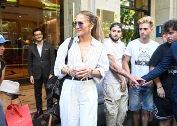 Jennifer Lopez, dressed in a striped blouse and white trousers, holds a coffee cup as she stands outside surrounded by fans and onlookers.