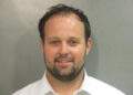 Josh Duggar in a mugshot photo wearing a white collared shirt, looking at the camera with a slight smile.