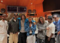 A group of seven people in a recording studio, with wood paneling and audio equipment in the background.