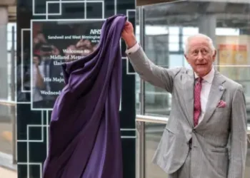 Richard Pohle/Pool via Reuters King Charles pulls a purple cloth from a black glass plaque saying NHS Sandwell and West Birmingham. Welcome to the Midland Metropolitan University Hospital. The plaque is partially obscured by the cloth. He is in a lobby area with glass screens behind him and looking to the left. He is wearing a grey suit and pink tie.
