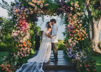 A couple kisses under a colorful floral arch at an outdoor wedding ceremony.