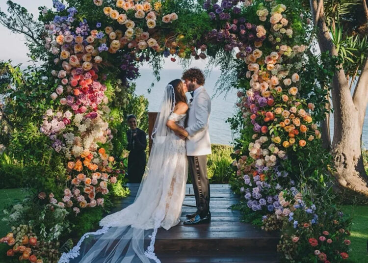 A couple kisses under a colorful floral arch at an outdoor wedding ceremony.