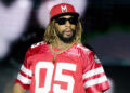 Lil Jon wearing a red sports jersey, sunglasses, and a red cap, with dreadlocks and a gold chain, performing on stage.