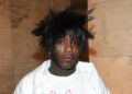 Lil Uzi Vert with spiky hair, facial piercings, and a white shirt, standing against a marble wall.