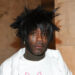 Lil Uzi Vert with spiky hair, facial piercings, and a white shirt, standing against a marble wall.