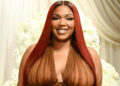Lizzo with long red hair, wearing a sheer brown outfit, smiling in front of a white floral backdrop.