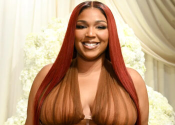 Lizzo with long red hair, wearing a sheer brown outfit, smiling in front of a white floral backdrop.