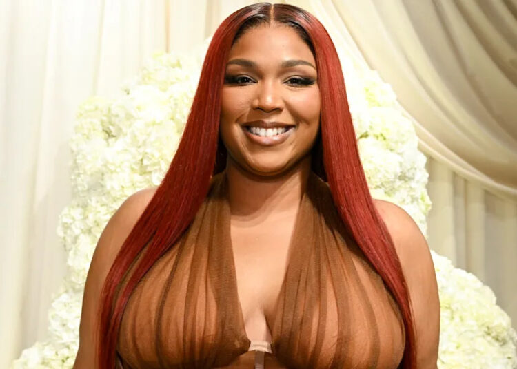 Lizzo with long red hair, wearing a sheer brown outfit, smiling in front of a white floral backdrop.
