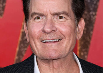 Charlie Sheen smiles on the red carpet, wearing a black textured blazer over a white shirt.