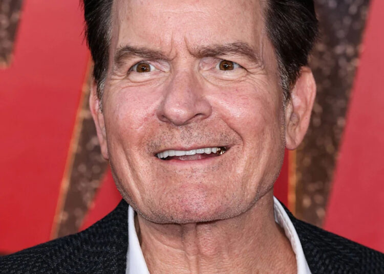 Charlie Sheen smiles on the red carpet, wearing a black textured blazer over a white shirt.