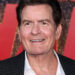 Charlie Sheen smiles on the red carpet, wearing a black textured blazer over a white shirt.