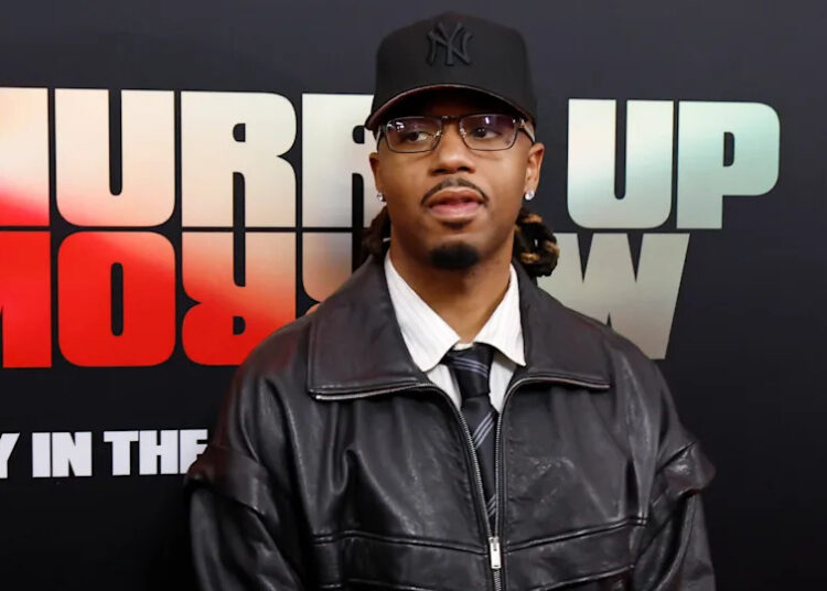 Metro Boomin wearing a black cap, glasses, leather jacket, and tie stands in front of a backdrop with text.