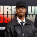Metro Boomin wearing a black cap, glasses, leather jacket, and tie stands in front of a backdrop with text.