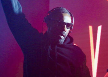 Idris Elba wearing headphones and sunglasses, he is a DJ at an event with bright red lights behind him
