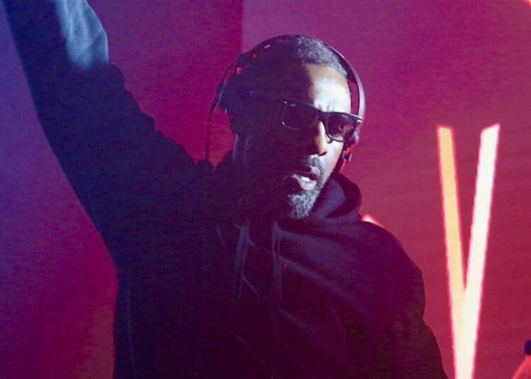 Idris Elba wearing headphones and sunglasses, he is a DJ at an event with bright red lights behind him