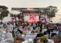 A crowd both standing and seated at tables watches a live music performance on an outdoor stage. A banner above the stage reads 'One Live! We support you first responders.'