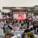 A crowd both standing and seated at tables watches a live music performance on an outdoor stage. A banner above the stage reads 'One Live! We support you first responders.'