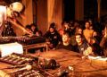 DJ on stage wearing a white t-shirt and cream cap. A large audience is watch. They are clapping and dancing to the performance. The stage has a lot of sound equipment on there not being used.