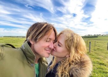 Nicole Kidman kisses Keith Urban on the cheek under a blue sky with white clouds.