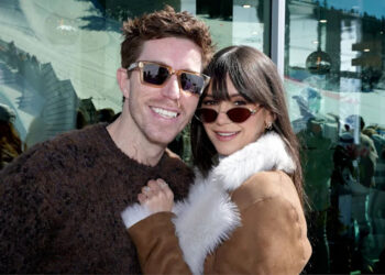 Shaun White and Nina Dobrev pose together outside a building, with snowy mountains reflected in the glass behind them.
