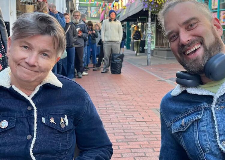 Zoe Cutting and Simon Smith queuing outside Resident Music in Kensington Gardens, Brighton and Hove. Both are wearing button up jackets; Zoe's is corduroy and Simon's is denim. They are both sitting in camp chairs.