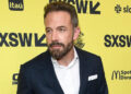 Ben Affleck at SXSW, wearing a dark suit with a white shirt, standing against a yellow background with event logos.