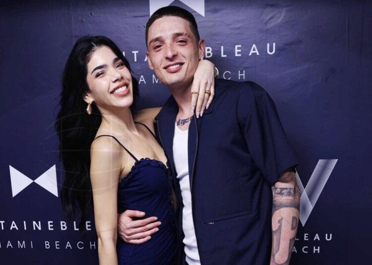 Kenia Os and Peso Pluma hug and pose in front of a step and repeat backdrop reading "Fontainebleau Miami Beach