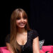 A woman with long hair in a black V-neck top, sits smiling on a pink couch with a black pillow