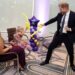 A man in a suit plays with a balloon near a young girl who is sitting down. Another woman who's also sitting is looking on.