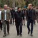 Three men walk next to each other talking with another group of people following behind.