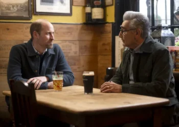 Prince William and Eugene Levy sitting at a bar table drinking beer