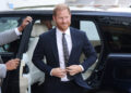 Prince Harry, wearing a navy blue suit and smiling, steps out of a black car