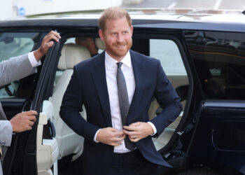 Prince Harry, wearing a navy blue suit and smiling, steps out of a black car