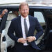 Prince Harry, wearing a navy blue suit and smiling, steps out of a black car