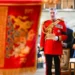 Finnbarr Webster Lieutenant General Sir Edward Smyth-Osbourne in a red military jacket and medals with HRH The Princess Royal. They are both holding a piece of paper and appear to be singing. In the foreground of the picture is a blurred red flag with a lion and a unicorn.