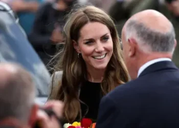 Reuters The Princess of Wales smiles and holds brightly coloured flowers whilst speaking to two men with their backs to the camera. She is wearing a black top and brown tweed-style suit and is surrounded by people and standing nearby to a large car.