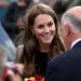 Reuters The Princess of Wales smiles and holds brightly coloured flowers whilst speaking to two men with their backs to the camera. She is wearing a black top and brown tweed-style suit and is surrounded by people and standing nearby to a large car.