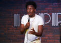 Comedian Kam Patterson wearing a white shirt, holding a microphone with a surprised expression, brick wall background with a logo.