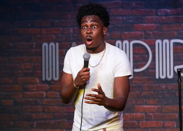 Comedian Kam Patterson wearing a white shirt, holding a microphone with a surprised expression, brick wall background with a logo.