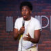 Comedian Kam Patterson wearing a white shirt, holding a microphone with a surprised expression, brick wall background with a logo.