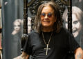 Ozzy Osbourne sitting on a throne-like chair, wearing sunglasses and a cross necklace, with a backdrop featuring his images.