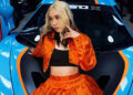 A woman with blonde hair wearing an orange outfit poses in front of a blue and orange sports car.